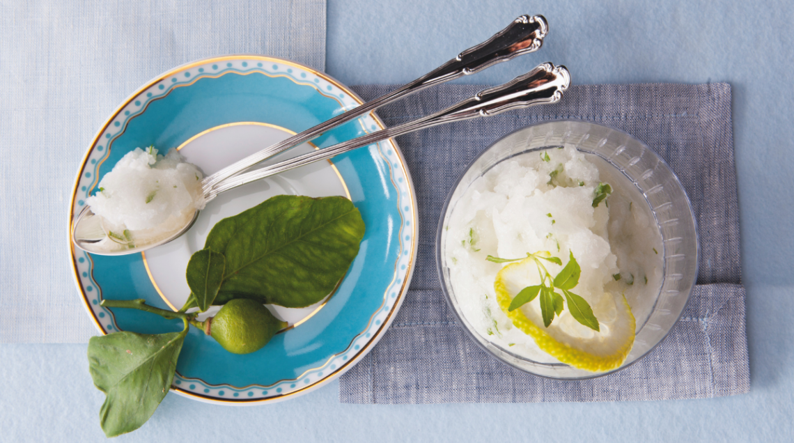 A sinistra un piatto bianco e azzurro con due cucchiaini lunghi e un po di sorbetto sopra e una foglia di limone, mentre sulla destra il sorbetto al basilico e limone in coppa trasparente