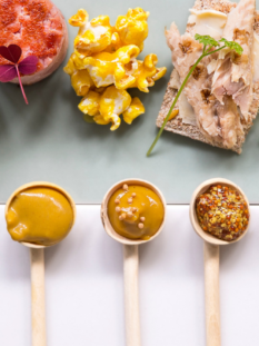 diversi cucchiaini contenenti salse a base di senape sono disposti orizzontalmente. Di fronte a ognuno c'è un ingredienti, come la tartare, un uovo sodo, un trancio di salmone ecc...