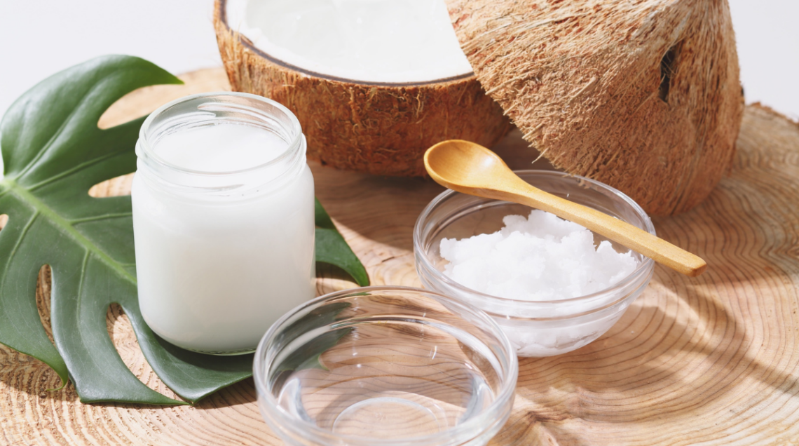 su una superficie di legno vi sono: un contenitore di vetro con olio di cocco, due coppette di vetro (una con olio di cocco e una con cocco rapé) e un noce di cocco sullo sfondo tagliata a metà
