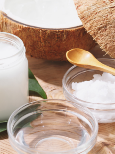 su una superficie di legno vi sono: un contenitore di vetro con olio di cocco, due coppette di vetro (una con olio di cocco e una con cocco rapé) e un noce di cocco sullo sfondo tagliata a metà