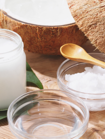 su una superficie di legno vi sono: un contenitore di vetro con olio di cocco, due coppette di vetro (una con olio di cocco e una con cocco rapé) e un noce di cocco sullo sfondo tagliata a metà