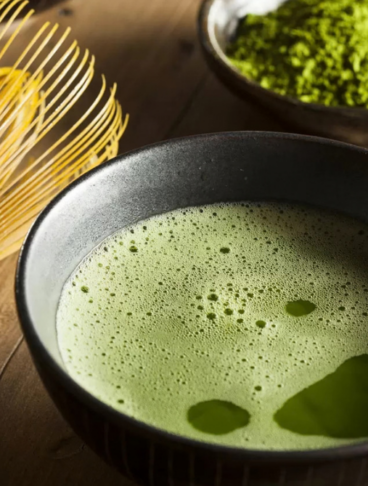 sullo sfondo, all'interno di una ciotolina, troviamo la polvere di tè matcha. In primo piano, all'interno di una ciotola rotonda, il tè matcha pronto (diluito con acqua) e l'apposito frustino di bambù che si utilizza per mescolarlo