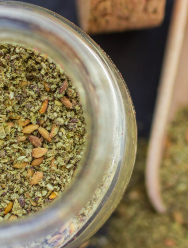 za'atar: nella foto una ciotola di vetro che contiene la miscela pronta per l'uso, con sullo sfondo, sfuocata, una piccola palettina dosatrice in legno con la miscela.