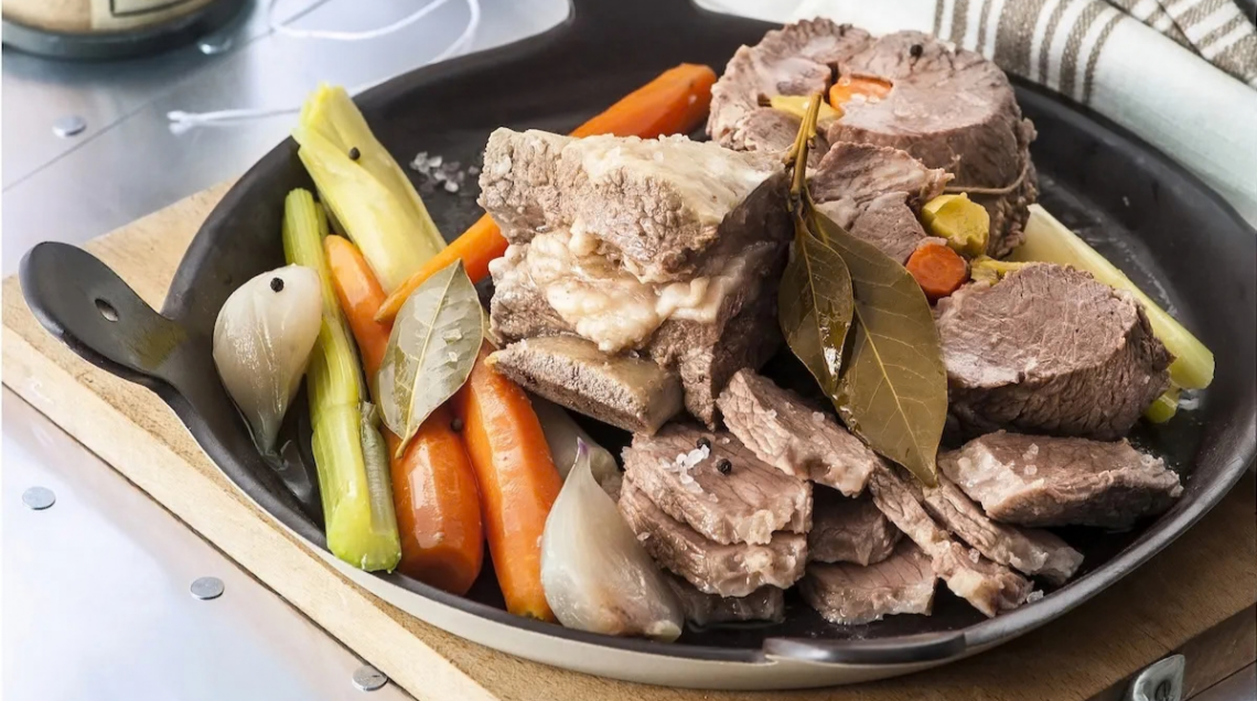 bollito di carne in una teglia nera rotonda con sedano e carote, su un tagliere di legno