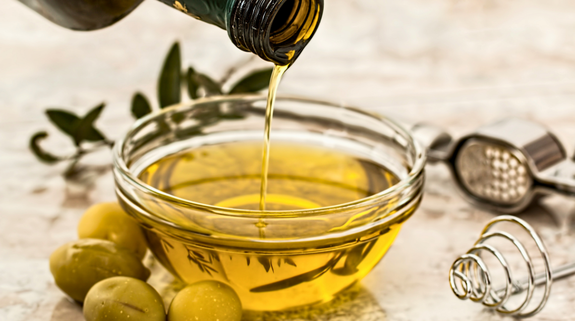 da una bottiglia viene versato dell'olio all'interno di una ciotolina di vetro, affianco alla quale vi sono delle olive