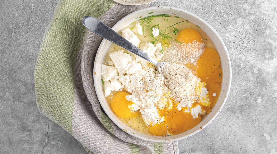 Inquadratura dall'alto (flat lay) che mostra la preparazione iniziale di una frittata. All'interno di una grande ciotola in ceramica chiara ci sono uova intere con i tuorli ben visibili, abbondante formaggio fresco sbriciolato (tipo caprino, feta o ricotta), una spolverata di formaggio grattugiato, pepe nero macinato e fili di erba cipollina sminuzzata. Un cucchiaio di metallo è parzialmente immerso negli ingredienti, pronto per amalgamare il tutto. Accanto alla ciotola si trovano una piccola tazza con altro formaggio grattugiato e un canovaccio nei toni del verde e del grigio, il tutto disposto su una superficie rustica in cemento o pietra grigia. 