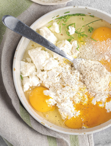 Inquadratura dall'alto (flat lay) che mostra la preparazione iniziale di una frittata. All'interno di una grande ciotola in ceramica chiara ci sono uova intere con i tuorli ben visibili, abbondante formaggio fresco sbriciolato (tipo caprino, feta o ricotta), una spolverata di formaggio grattugiato, pepe nero macinato e fili di erba cipollina sminuzzata. Un cucchiaio di metallo è parzialmente immerso negli ingredienti, pronto per amalgamare il tutto. Accanto alla ciotola si trovano una piccola tazza con altro formaggio grattugiato e un canovaccio nei toni del verde e del grigio, il tutto disposto su una superficie rustica in cemento o pietra grigia. 
