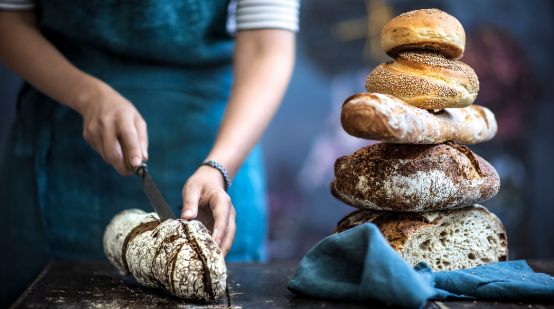 diversi pani impilati uno sull'altro e, a sinistra, una signora che taglia un pezzo di pane
