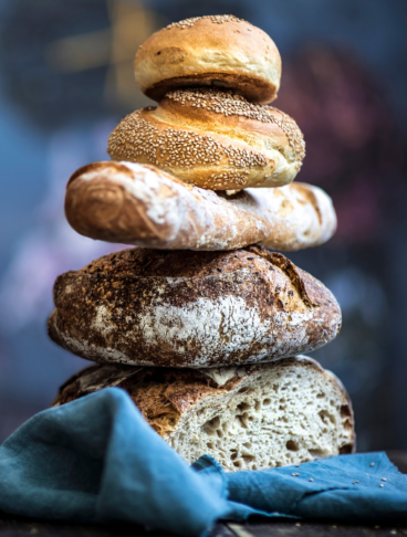 diversi pani impilati uno sull'altro e, a sinistra, una signora che taglia un pezzo di pane