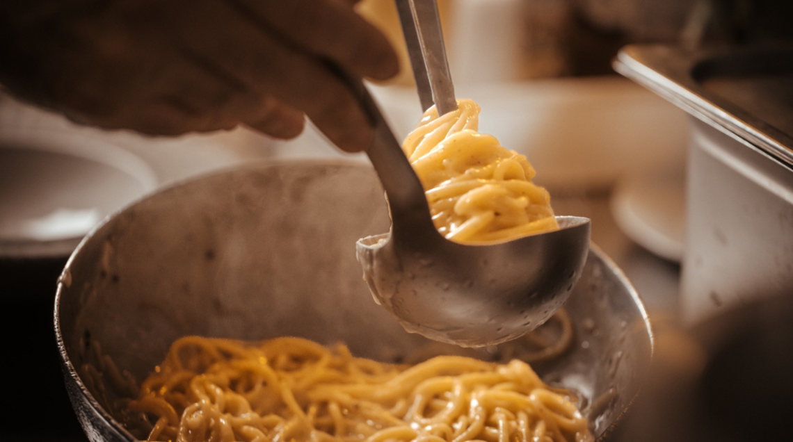 Con l'aiuto di una pinza da cucina in metallo, la pasta della Trattoria della Stampa viene sollevata e arrotolata a nido direttamente dentro un mestolo d'acciaio, prelevandola da una padella d'alluminio visibile in primo piano