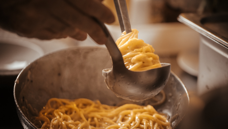 Con l'aiuto di una pinza da cucina in metallo, la pasta della Trattoria della Stampa viene sollevata e arrotolata a nido direttamente dentro un mestolo d'acciaio, prelevandola da una padella d'alluminio visibile in primo piano