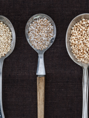 diversi tipi di cucchiai appoggiati su uno sfondo nero e riempiti con quinoa, amaranto e grano saraceno